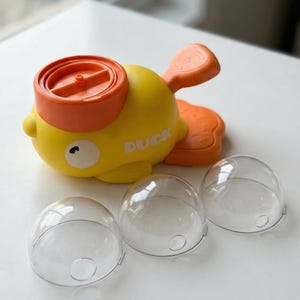 May include: A yellow and orange toy duck with the word "DUCK" printed on its side. The duck has a propeller on its head and a tail fin. Three clear, dome-shaped plastic containers are in front of the duck.