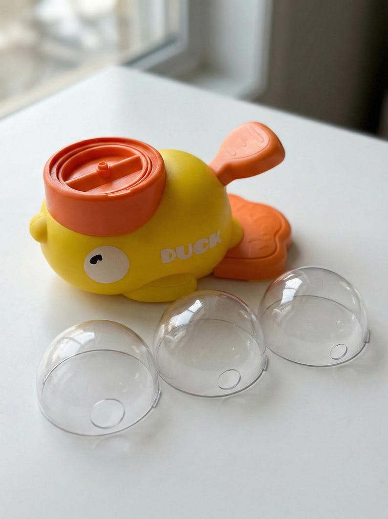May include: A yellow and orange toy duck with the word "DUCK" printed on its side. The duck has a propeller on its head and a tail fin. Three clear, dome-shaped plastic containers are in front of the duck.