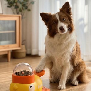 May include: A brown and white dog sits next to a yellow and orange dog toy with a clear dome filled with dog food. The toy has a duck design and the dog is resting its paw on the toy.