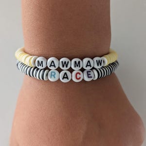 May include: Two stacked bracelets made of flat circular beads. The top bracelet is pale yellow with white letter beads spelling MAWMAW. The bottom bracelet features black and white stripes with white letter beads spelling RACE in multicolored letters.