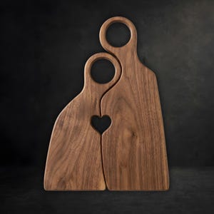 Puede incluir: Dos tablas de cortar de madera de color marrón oscuro que encajan entre sí con vetas naturales. Cada tabla tiene un mango circular en la parte superior y, al unirlas, forman un pequeño recorte en forma de corazón en el centro.