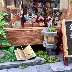 May include: Miniature Japanese shop scene with a sign that reads "New Products Released" in Japanese. The scene includes a stone lantern, a small open book, and various miniature items displayed on wooden shelves.
