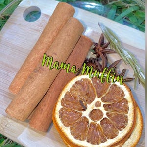 May include: Three brown cinnamon sticks, two dark brown star anise, a bundle of dried green herbs, and three dried orange slices arranged on a light wood cutting board. The text Mama Muffin is printed in green across the center of the items.