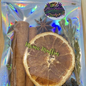 May include: A holographic pouch containing dried botanicals including two cinnamon sticks, a star anise, a dried orange slice, sprigs of rosemary, and cloves. The pouch features a circular logo with the text Mama Muffin's Herbal Care & Wellness LLC.