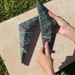 Puede incluir: Dos sujetalibros de mármol verde oscuro con una forma geométrica triangular facetada. La piedra presenta vetas blancas prominentes en toda la superficie, creando un patrón jaspeado distintivo en cada una de las dos piezas decorativas de piedra.