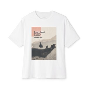 May include: A white short-sleeved t-shirt featuring a rectangular graphic print on the chest. The print displays a seagull standing near an outstretched hand over a body of water, with the text Everything I want, wants me more. in black.