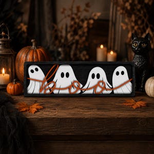 May include: A rectangular black wooden sign featuring four white ghost illustrations with black eyes and mouths. The word "boo" is written across the ghosts in a continuous orange cursive script. The sign has a dark frame and a smooth finish.