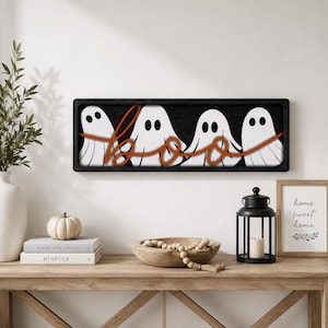 May include: A rectangular black wooden wall sign featuring four white ghost figures with black eyes and mouths. The word "boo" is written in a cursive, orange-colored script that overlays the ghosts, spanning across the entire width of the sign.