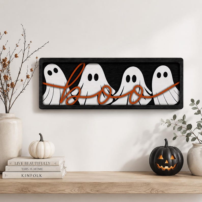 May include: A rectangular wall sign with a black frame featuring four white ghost figures with black eyes and mouths. The word "boo" is written in a cursive, orange-colored script that overlays the ghosts across the center of the sign.