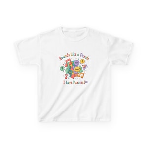May include: A white short-sleeved t-shirt featuring a colourful graphic of interlocking puzzle pieces, musical notes, and speech bubbles with the text "Sounds Like a Puzzle" and "I Love Puzzles!" printed in a multi-coloured, rounded font style.