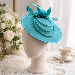 Puede incluir: Un tocado de color turquesa con un ala circular ancha y un acabado liso y estructurado. La parte superior está adornada con tres bucles circulares concéntricos y un grupo de plumas y pétalos de tela a juego dispuestos en un diseño floral.