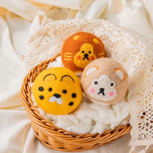 May include: Three felt animal-faced balls in a woven basket. The balls feature a lion, a cheetah, and a bear design. The lion is orange, the cheetah is yellow, and the bear is tan. The basket sits on a white, textured surface.