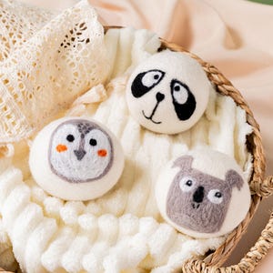 May include: Three white wool dryer balls, each with a different animal face: a panda, an owl, and a koala. The panda has black markings, the owl has orange cheeks, and the koala is grey. They are in a woven basket on a white, textured surface.