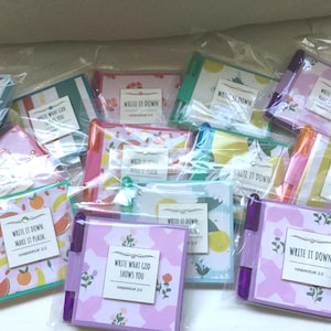 May include: A collection of colorful notebooks and markers, each packaged in clear plastic. The notebooks feature floral and fruit patterns, with text that reads "WRITE IT DOWN" and "WRITE WHAT GOD SHOWS YOU" from Habakkuk 2:2. Markers are orange, purple, and green.