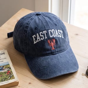 Può includere: Un cappellino da baseball in denim blu con la scritta ricamata in bianco EAST COAST sopra una piccola grafica di un'aragosta rossa. Il cappellino presenta una visiera curva e una corona strutturata con occhielli di ventilazione.