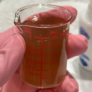 May include: A clear glass beaker with red measurement markings, filled with a brown liquid. The beaker is held by a person wearing pink gloves. The markings include ounces and milliliters.