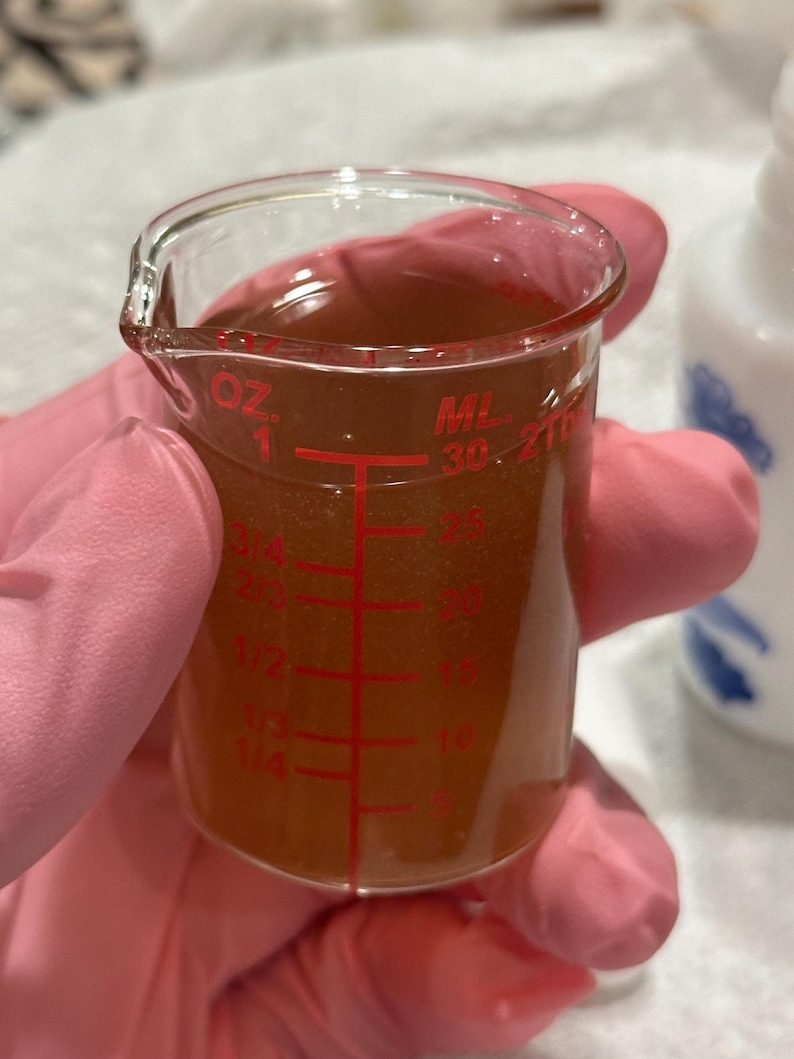 May include: A clear glass beaker with red measurement markings, filled with a brown liquid. The beaker is held by a person wearing pink gloves. The markings include ounces and milliliters.