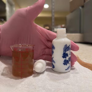 May include: A white ceramic bottle with blue floral designs, a clear glass beaker with a brown liquid, and a white dropper are arranged on a white surface. The bottle is cylindrical, and the beaker has red measurement markings. The dropper is white.