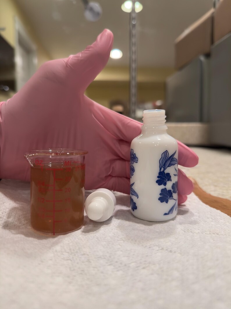 May include: A white ceramic bottle with blue floral designs, a clear glass beaker with a brown liquid, and a white dropper are arranged on a white surface. The bottle is cylindrical, and the beaker has red measurement markings. The dropper is white.