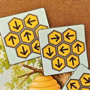 May include: Three square game tiles featuring a light blue background and a cluster of yellow hexagonal cells. Each hexagon contains a black arrow pointing in various directions, including up, down, left, and right, arranged in a honeycomb pattern.