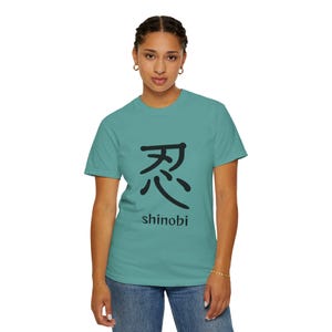 以下が含まれることがあります： 黒い日本語の漢字と「shinobi」の文字が入った、ターコイズ色のTシャツ。Tシャツはクルーネックです。その人は青いジーンズを履いています。