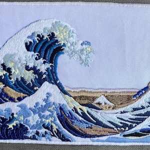 May include: A rectangular fabric patch featuring a detailed embroidery of a large crashing ocean wave in shades of deep blue, light blue, and white, with a small mountain visible in the background against a plain light-colored fabric background.