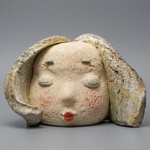 Könnte beinhalten: Eine Keramikskulptur eines Gesichts mit geschlossenen Augen, roten Wangen und roten Lippen. Das Haar ist strukturiert und umgibt das Gesicht. Die Skulptur ist hellbeige mit dunkleren Akzenten.