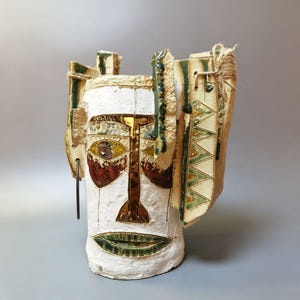 Könnte beinhalten: Handgefertigte Keramikvase mit Gesichtsdesign. Die Vase hat eine weiße Basis mit braunen, grünen und goldenen Akzenten. Das Gesicht hat ein Auge, eine Nase und einen Mund. Dekorative Elemente sind Seil und strukturierte Paneele. Die Vase ist etwa 30 cm hoch.