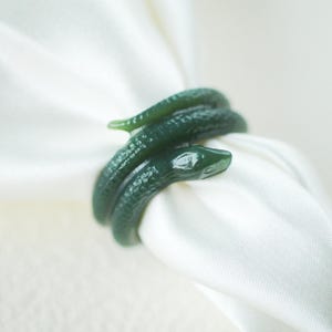 May include: A dark green, snake-shaped napkin ring with a textured, scale-like surface, coiled around a folded white fabric napkin. The snake design features a clearly defined head and a slender, tapering tail that wraps around the cloth.