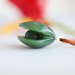 May include: A dark green pendant shaped like an open clam shell with a smooth, round dark green bead nestled inside. The pendant features a ribbed texture on the exterior shell and is attached to a brown cord by a silver-toned metal jump ring.