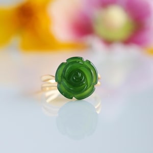 May include: A gold ring featuring a carved green jade rose. The rose is the focal point, with detailed petals. The ring's band is a simple, polished gold. The background is blurred with soft pink, yellow, and green hues.