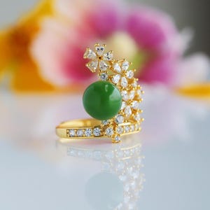 Op de afbeelding: Een gouden ring met een grote, ronde, groene edelsteen. De ring is versierd met kleine, heldere edelstenen en bloemmotieven. Het ontwerp staat tegen een zachte, vage achtergrond van roze en geel.
