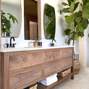 May include: A modern bathroom vanity with a light wood finish, featuring multiple drawers and a lower shelf with woven baskets. The countertop is white, with black faucets and oval mirrors with gold frames. A large potted plant adds a touch of greenery.