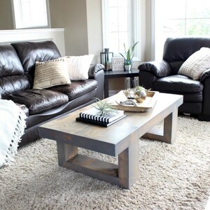 May include: A living room scene featuring a dark brown leather sofa and armchair, a light gray wooden coffee table, and a cream-colored rug. Decorative pillows and a white throw blanket add texture. A small black side table holds a plant.