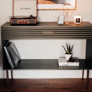Puede incluir: Una consola gris oscuro con un tocadiscos Victrola vintage, discos de vinilo y objetos decorativos. La mesa tiene un frente de cajón acanalado, herrajes dorados y patas de horquilla. Una pequeña planta en maceta y libros están en el estante inferior.