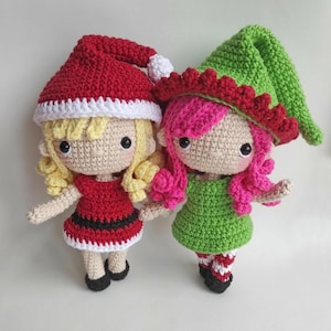 May include: Two handmade crochet dolls. One doll has blonde hair, a red dress with a black belt, and a red Santa hat with a white trim. The other doll has pink hair, a green dress, and a green pointed hat with a red ruffled brim.