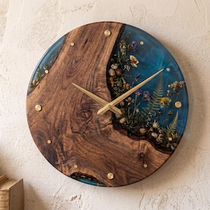 Op de afbeelding: Ronde houten wandklok met een natuurlijk houtnerfdesign en een blauwe harsinleg. De wijzerplaat heeft gedroogde bloemen en kleine stenen ingebed in de hars. Goudkleurige wijzers en messing accenten maken het ontwerp compleet.