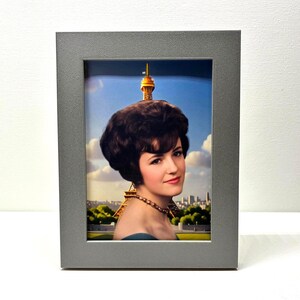 May include: A rectangular gray picture frame containing a portrait of a person with dark, voluminous hair and a beaded necklace. The background of the portrait features the Eiffel Tower against a blue sky with white clouds and a city skyline.