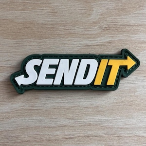 May include: A rectangular patch with the words "SEND IT" in white block letters. The word "IT" is in yellow with a right-pointing arrow. The patch has a dark green border and a light brown background.