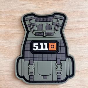 May include: A patch in the shape of a tactical vest, featuring the number "5.11" in white text on a black background. The vest is primarily gray and green with black accents, and includes details like straps and buckles.