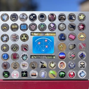 May include: A silver-colored game board with numerous circular tokens featuring various designs, including skulls, dice, and a radar screen. The board has the text "FNG" and "S.S.D.D." printed on it. The board is set on a red surface.