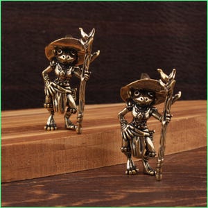 May include: Two bronze-colored frog figurines, each wearing a hat and holding a staff. The frog-like figures have detailed features and are posed on a wooden surface. The background is a dark brown color.