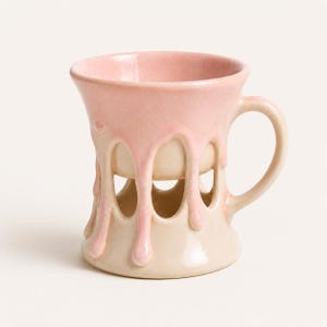Puede incluir: Una taza de cerámica con un diseño bicolor que presenta un esmalte rosa claro brillante que gotea sobre una base de color crema. La taza tiene un borde ancho, un asa curva y una serie de aberturas arqueadas en la parte inferior del cuerpo.