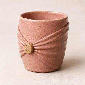 May include: A dusty rose ceramic cup with a speckled texture. The exterior features a raised, pleated design that gathers toward a central, circular tan button-like accent on the front, creating a draped fabric appearance on the vessel surface.