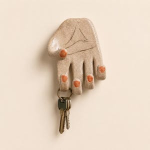 Pode incluir: Um gancho de parede de cerâmica em forma de mão com acabamento bege salpicado e unhas pintadas de laranja. A mão é moldada com dedos curvados para formar ganchos, e um porta-chaves de metal com três chaves prateadas está pendurado no dedo médio.