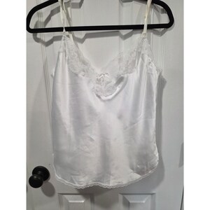 Pode incluir: Uma camisola de cetim branco com decote em V adornado com delicada renda branca e um pequeno laço central. A peça possui alças finas ajustáveis e uma bainha de renda recortada ao longo da borda inferior do tecido.
