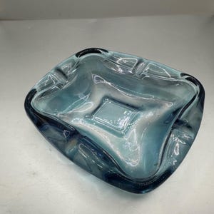 May include: A square, teal-colored ashtray with a rounded rim. The ashtray has a slightly textured surface and a dark blue border. The ashtray has four indentations for resting cigarettes.
