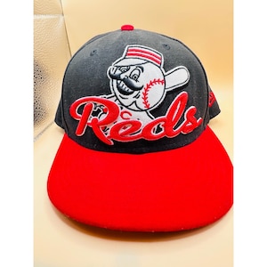 Könnte beinhalten: Baseballkappe mit rotem Schirm und schwarzer Krone. Die Kappe zeigt das Wort "Reds" in roter Schrift und eine Cartoon-Baseballfigur. Der obere Knopf ist rot.