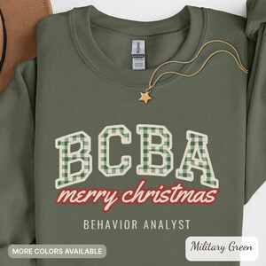 May include: An olive green crewneck sweatshirt featuring the text BCBA in a white and green gingham pattern. Below, the words merry christmas appear in red script, followed by the text BEHAVIOR ANALYST in white capital letters.