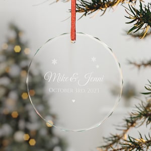 Puede incluir: Un adorno de cristal transparente circular con bordes facetados, colgado de una cinta roja. Está grabado con los nombres Mike & Jenni, la fecha OCTOBER 3RD, 2023, pequeños iconos de copos de nieve y un pequeño corazón.